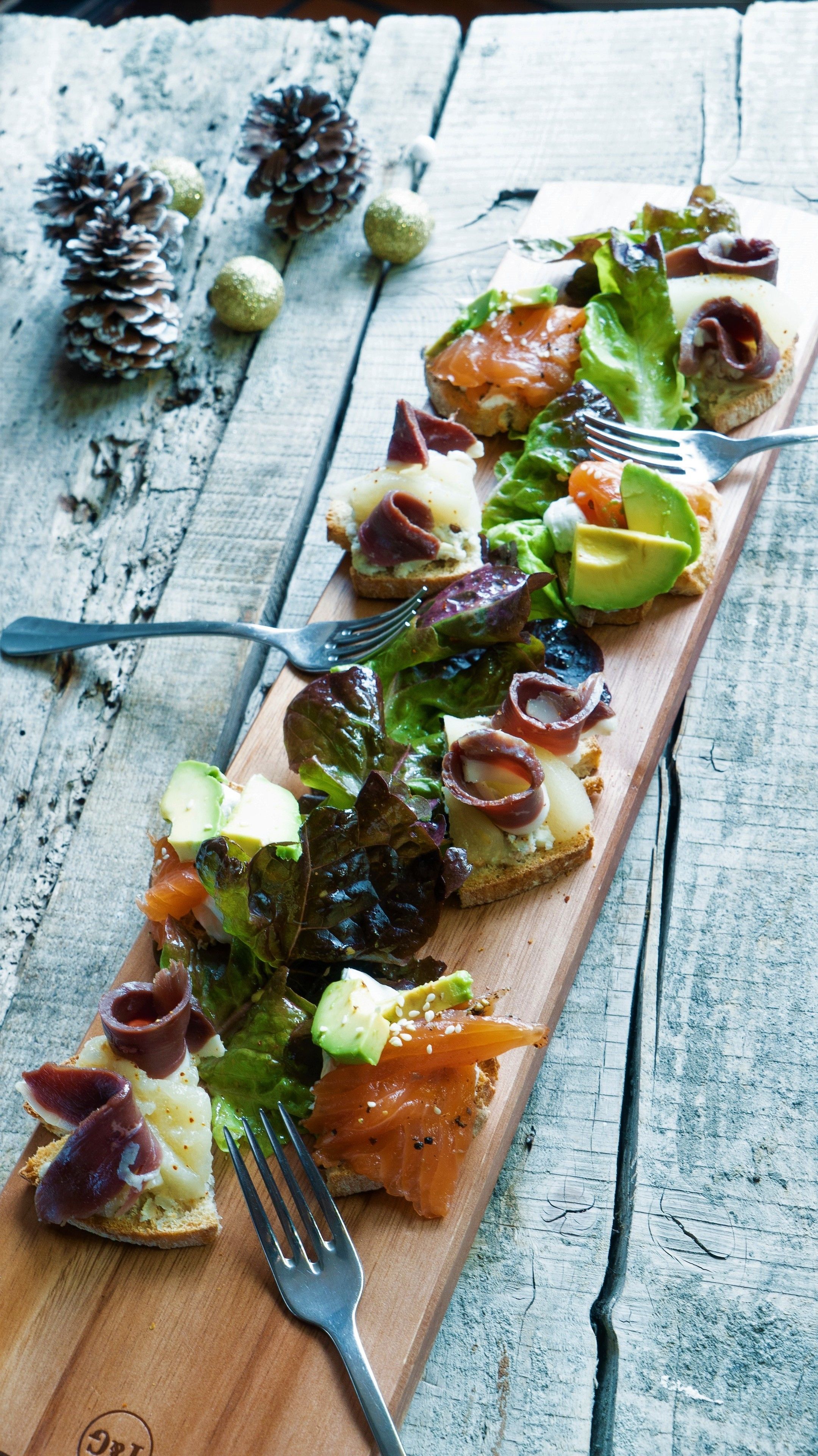 A wooden serving board with a variety of appetizers including smoked salmon, avocado, and greens, accompanied by a fork.