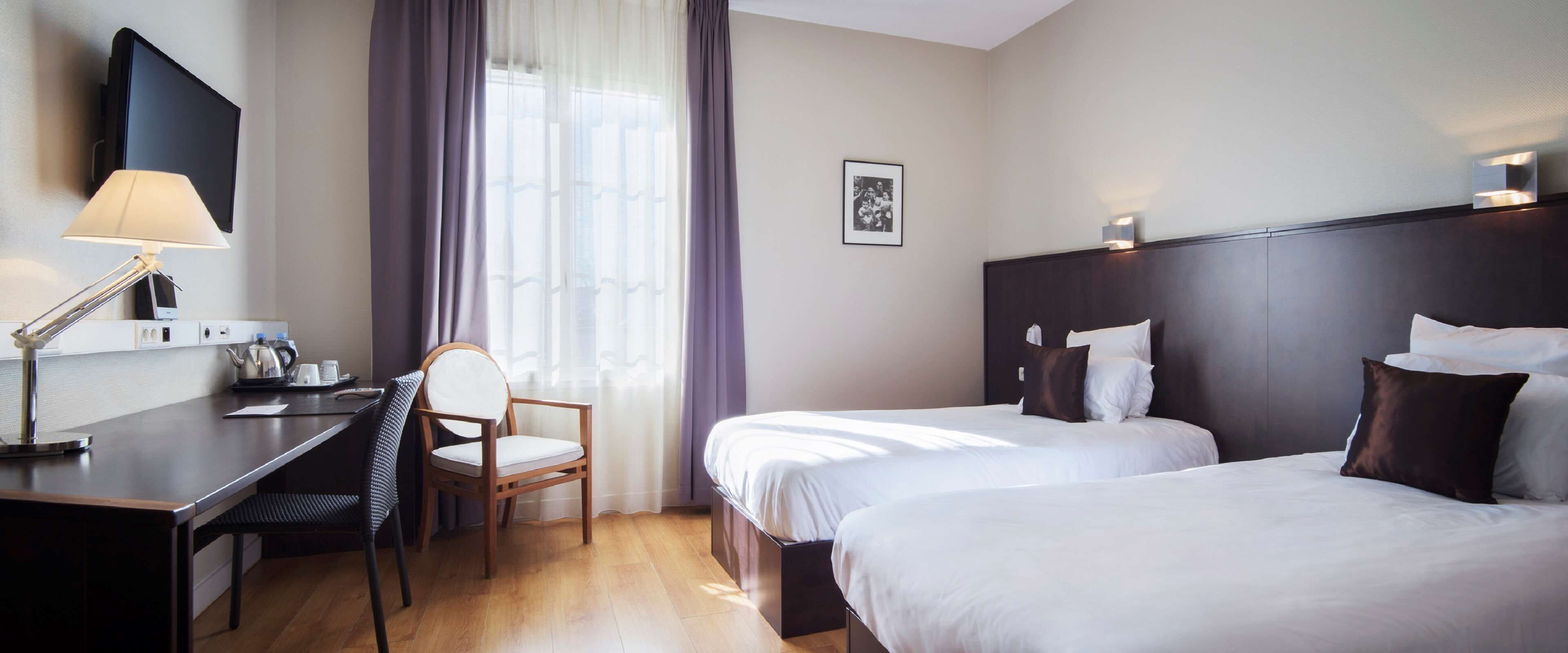 Une chambre d’hôtel équipée d’un bureau, d’une chaise, de deux lits, d’une télévision et d’une fenêtre avec des rideaux.