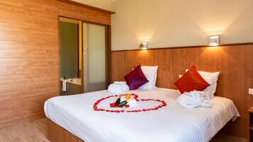 A hotel room with a wooden floor, a bed with white bedding, red and white pillows, a heart-shaped arrangement of rose petals, and a bottle of champagne.