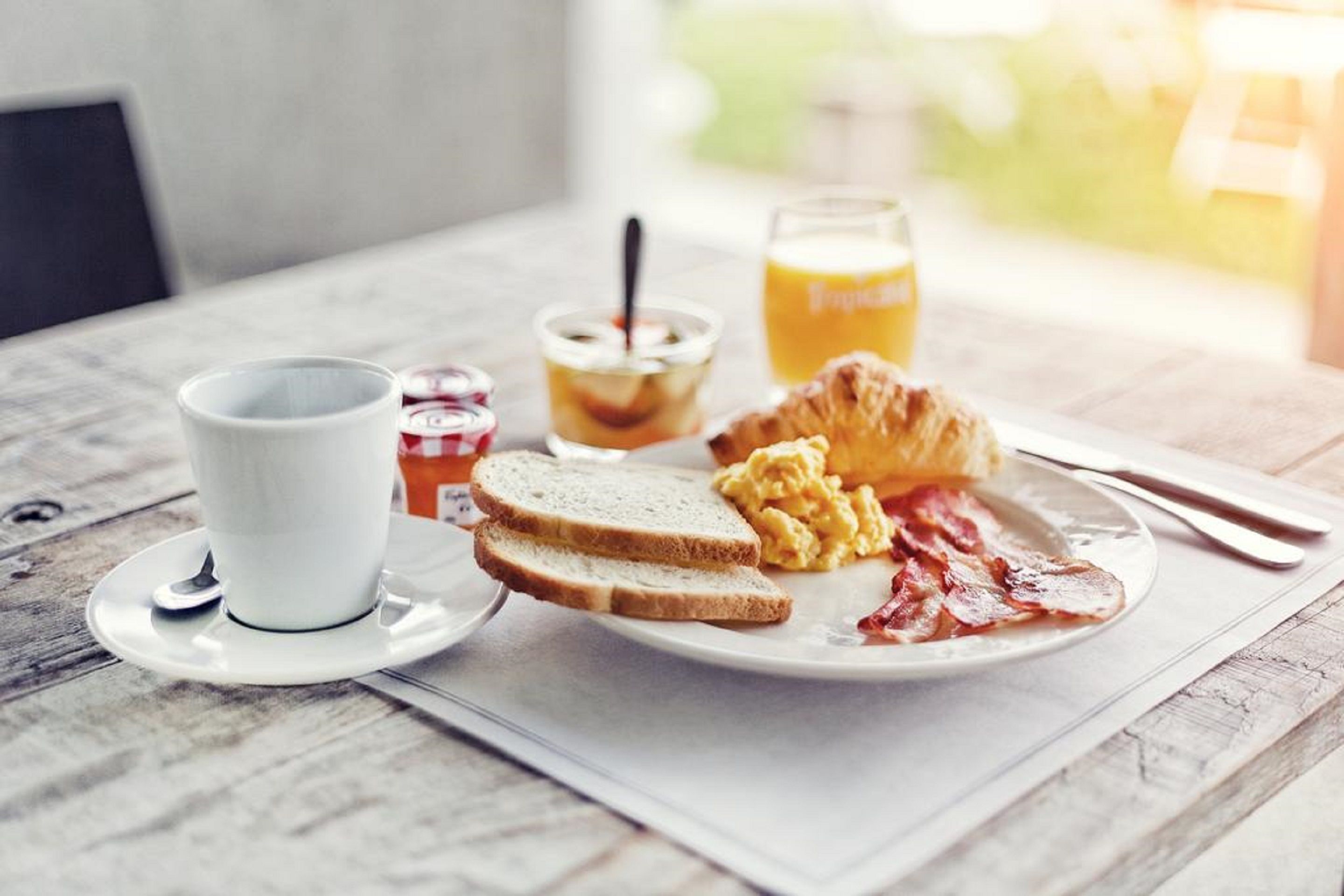 Aamiaispöytä, jossa on paahtoleipää, munakokkelia, pekonia, kuppi kahvia, lasi appelsiinimehua ja pieni purkki hilloa puupöydällä.
