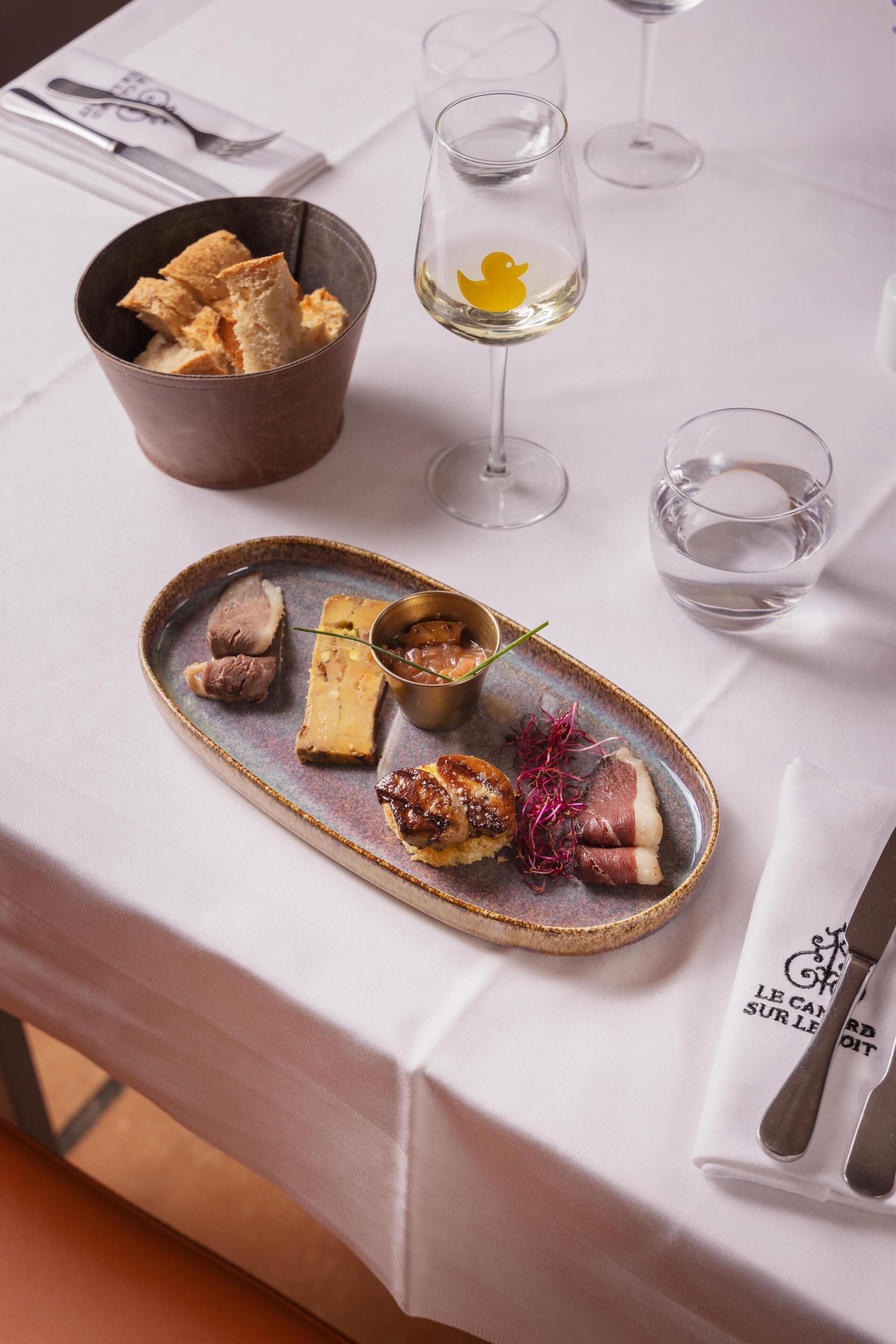 A dining table with a plate of food, a bowl of bread, and two glasses of white wine.