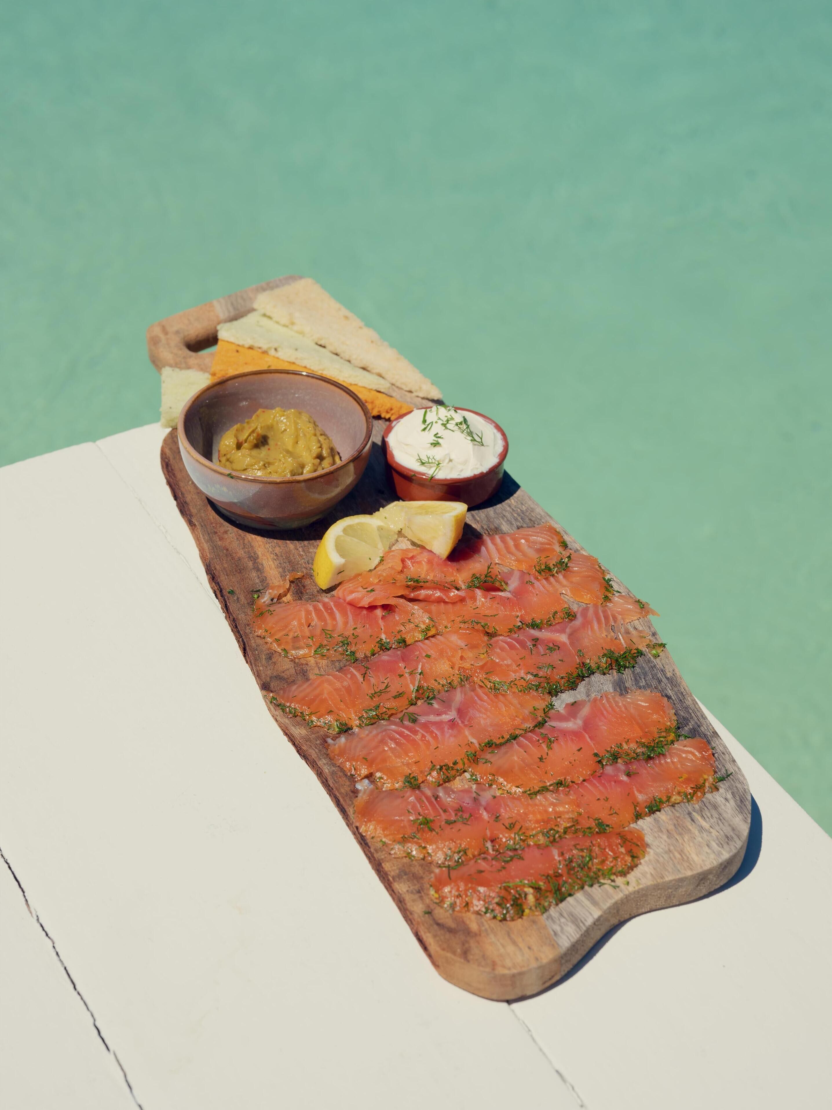 Una tabla de madera con salmón ahumado, un recipiente de mostaza y un tazón pequeño de queso crema decorado con hierbas.