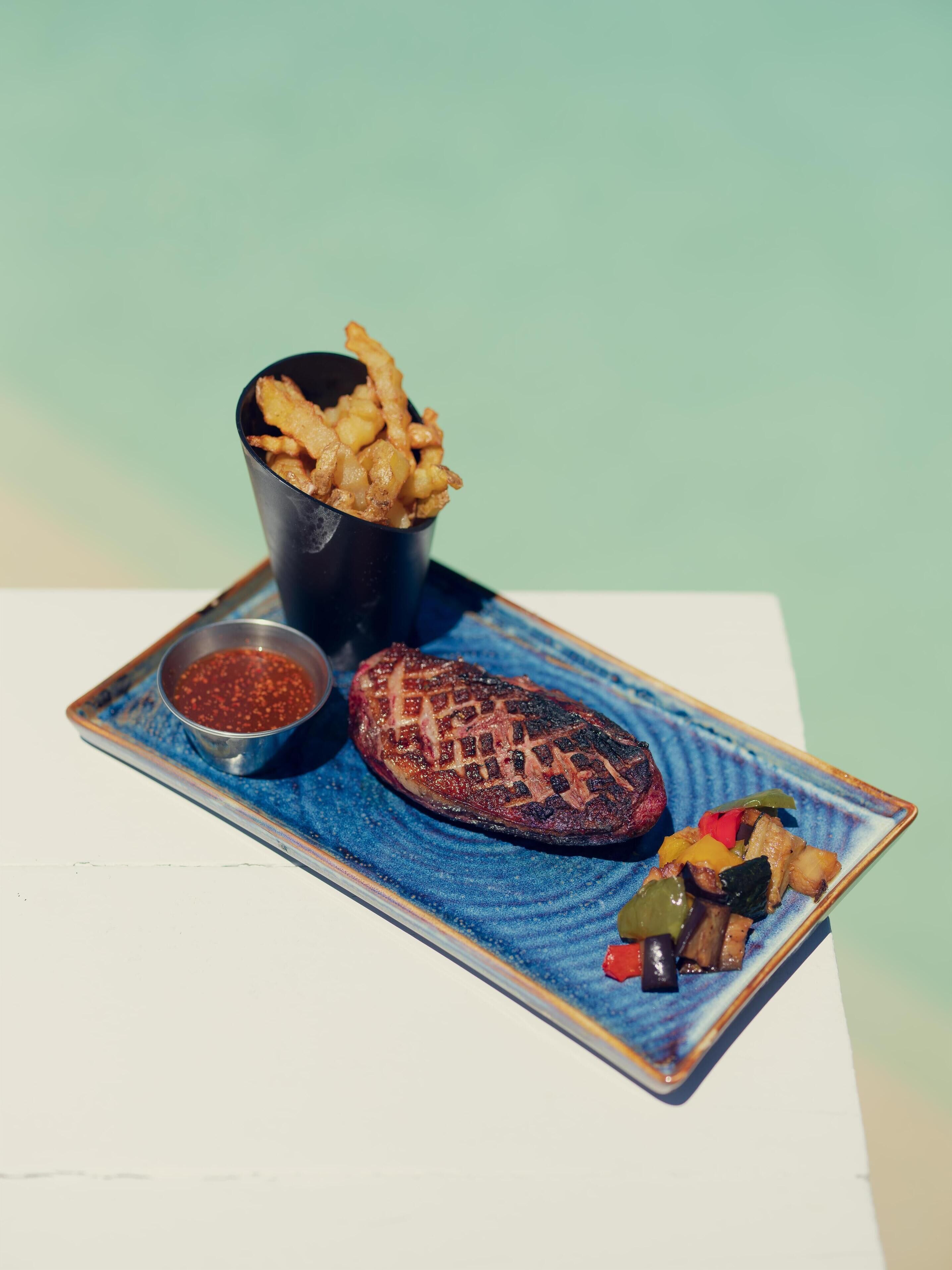 A plate with a grilled steak, a side of fries, and a small bowl of sauce.