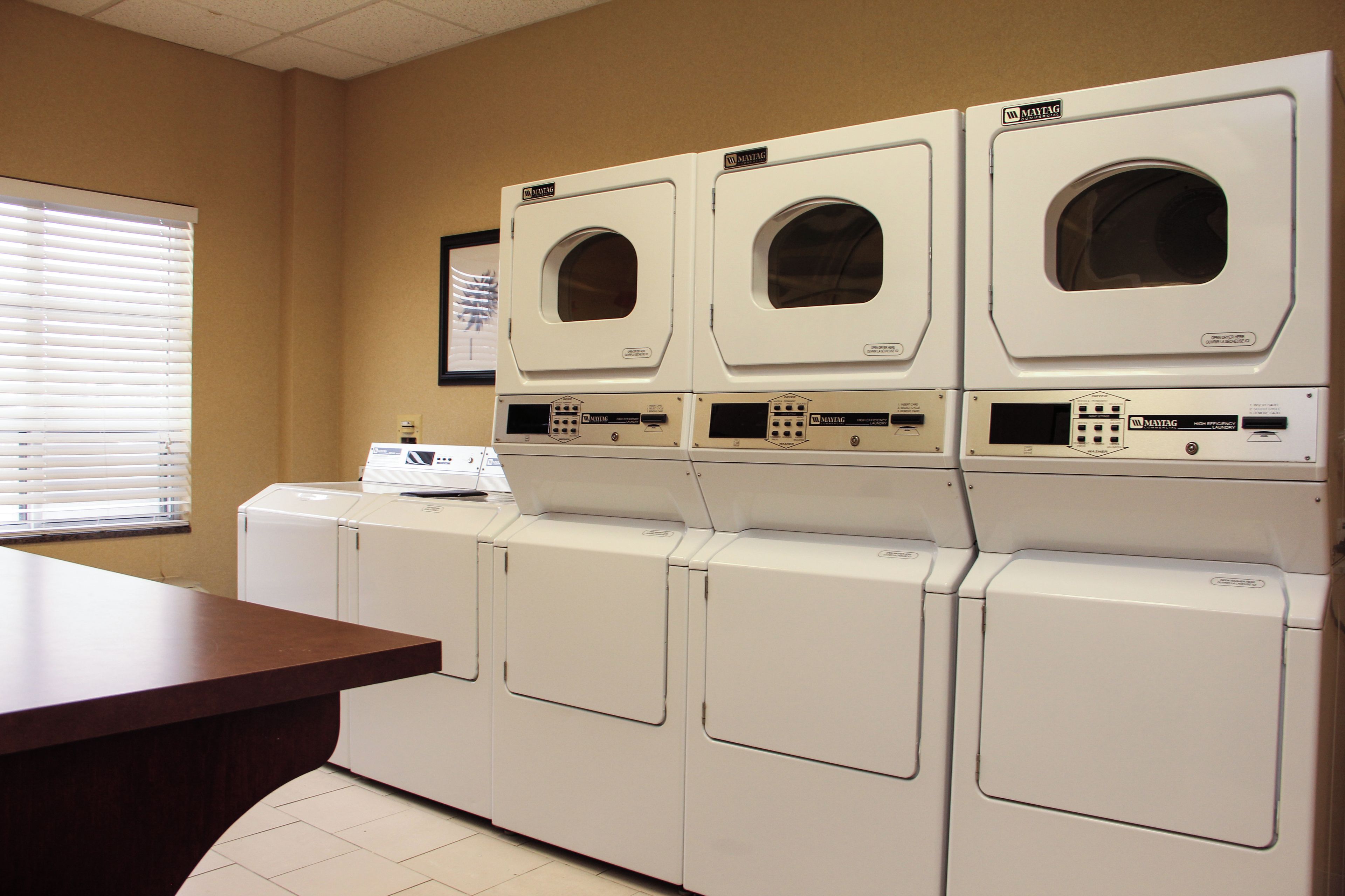A laundry room with multiple white Maytag washers and dryers, a wooden counter, and a window with blinds.