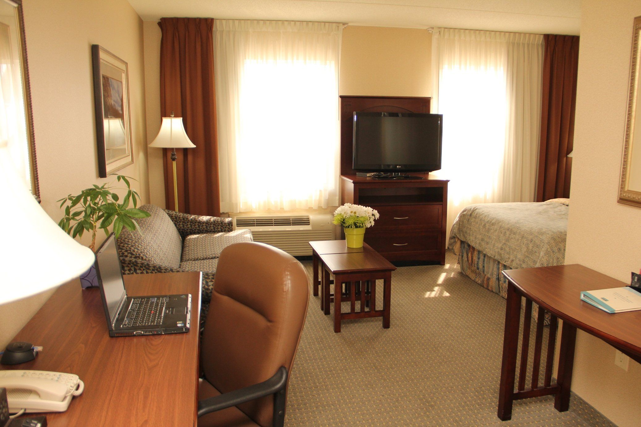 A hotel room with a desk, chair, sofa, TV, and a small table with flowers.