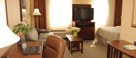 A hotel room with a desk, chair, sofa, TV, and a small table with flowers.