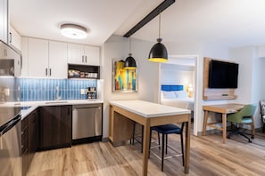 A modern kitchen with a built-in dishwasher, a dining area with a wooden table and bar stools, a TV mounted on the wall, and a colorful artwork on the wall. - Staybridge Suites London by IHG (London)