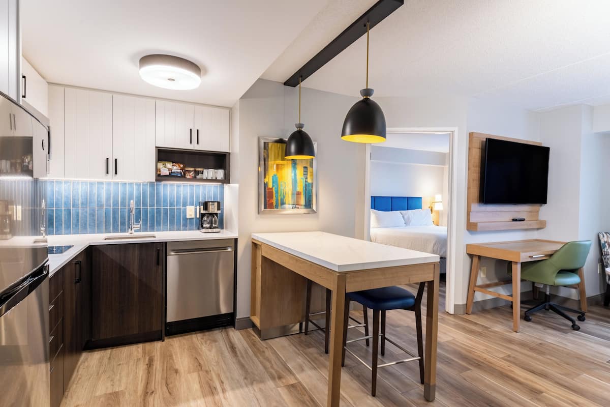 A modern kitchen with a built-in dishwasher, a dining area with a wooden table and bar stools, a TV mounted on the wall, and a colorful artwork on the wall.