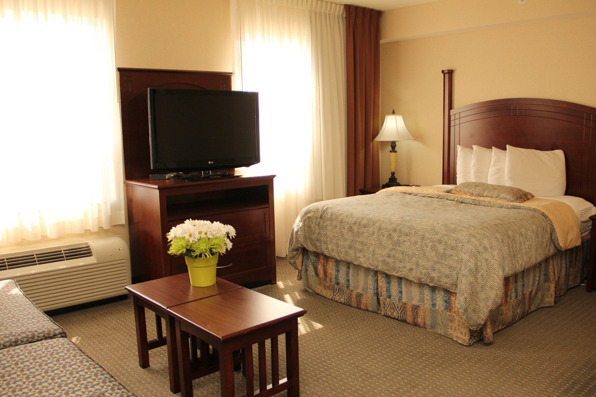 Quarto de hotel com cama, televisão sobre suporte de madeira, pequena mesa com vaso de flores, luminária e ar condicionado.
