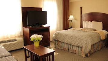 Quarto de hotel com cama, televisão sobre suporte de madeira, pequena mesa com vaso de flores, luminária e ar condicionado.