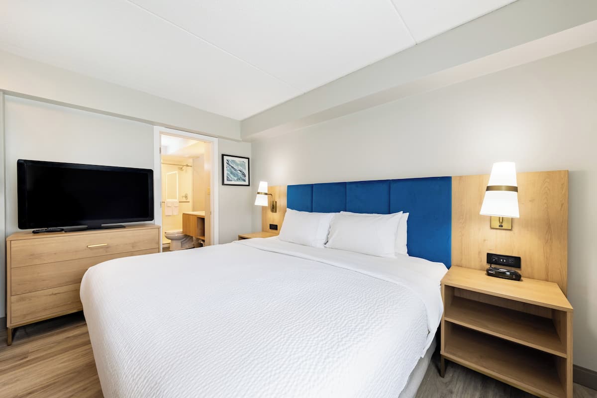 A hotel room with a large bed, a TV on a wooden dresser, a nightstand with a lamp, and a bathroom visible through an open door.