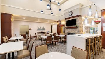A hotel dining area with multiple tables and chairs, a central island with bar stools, a fireplace, and a television mounted on the wall.