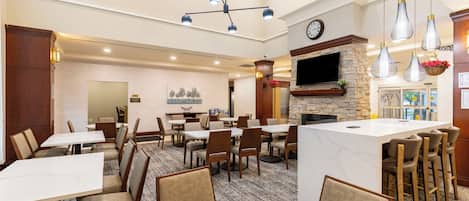 Comedor de hotel con varias mesas y sillas, una isla central con taburetes, una chimenea y un televisor montado en la pared.