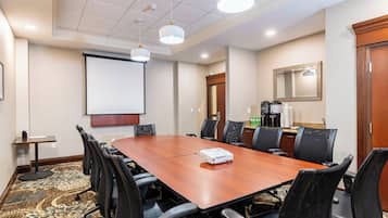 Un salón de conferencias con una mesa de madera, sillas negras, un proyector, una pantalla y un espacio para café.