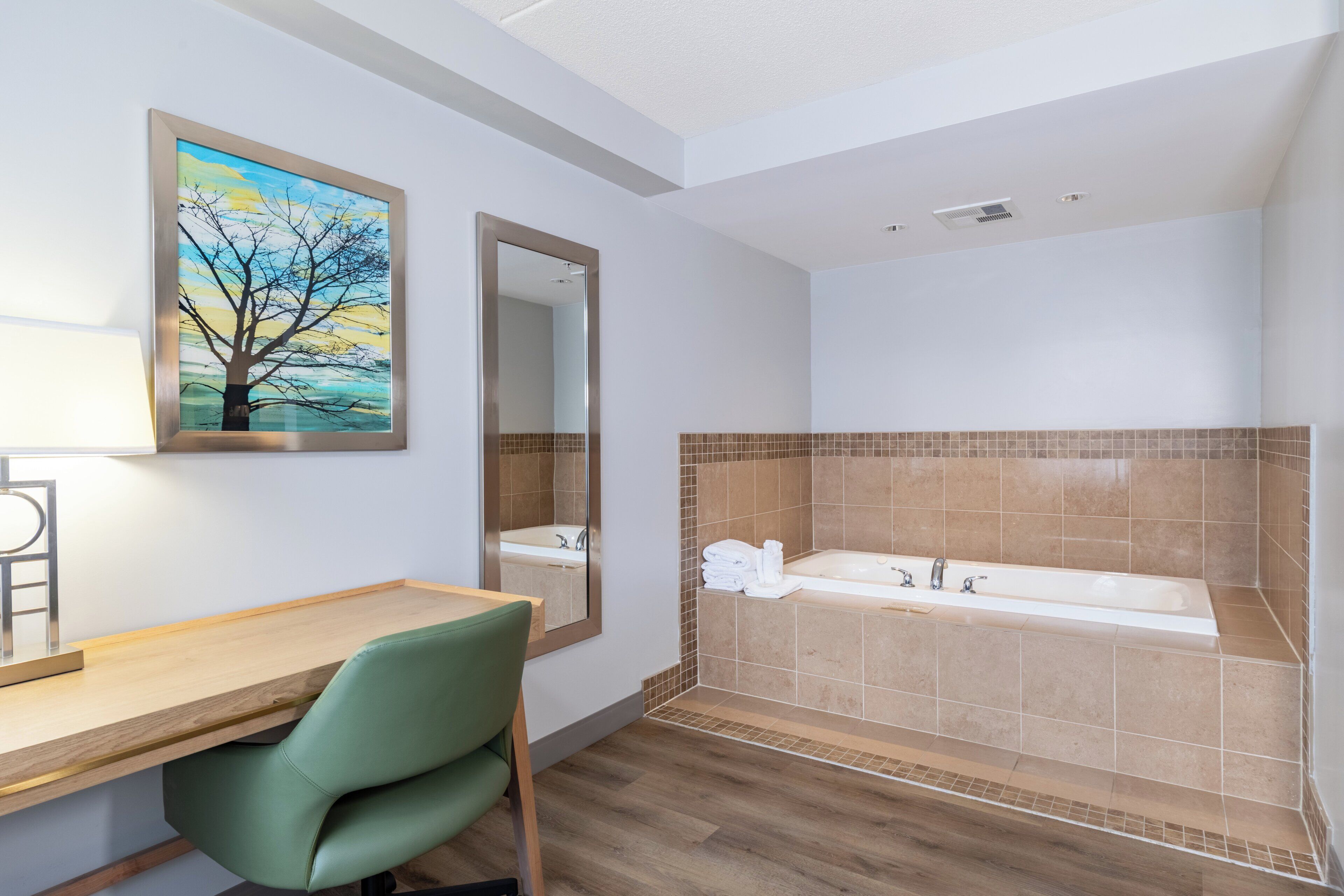 A modern bathroom with a wooden vanity, a green chair, a large bathtub, and a painting of a tree.
