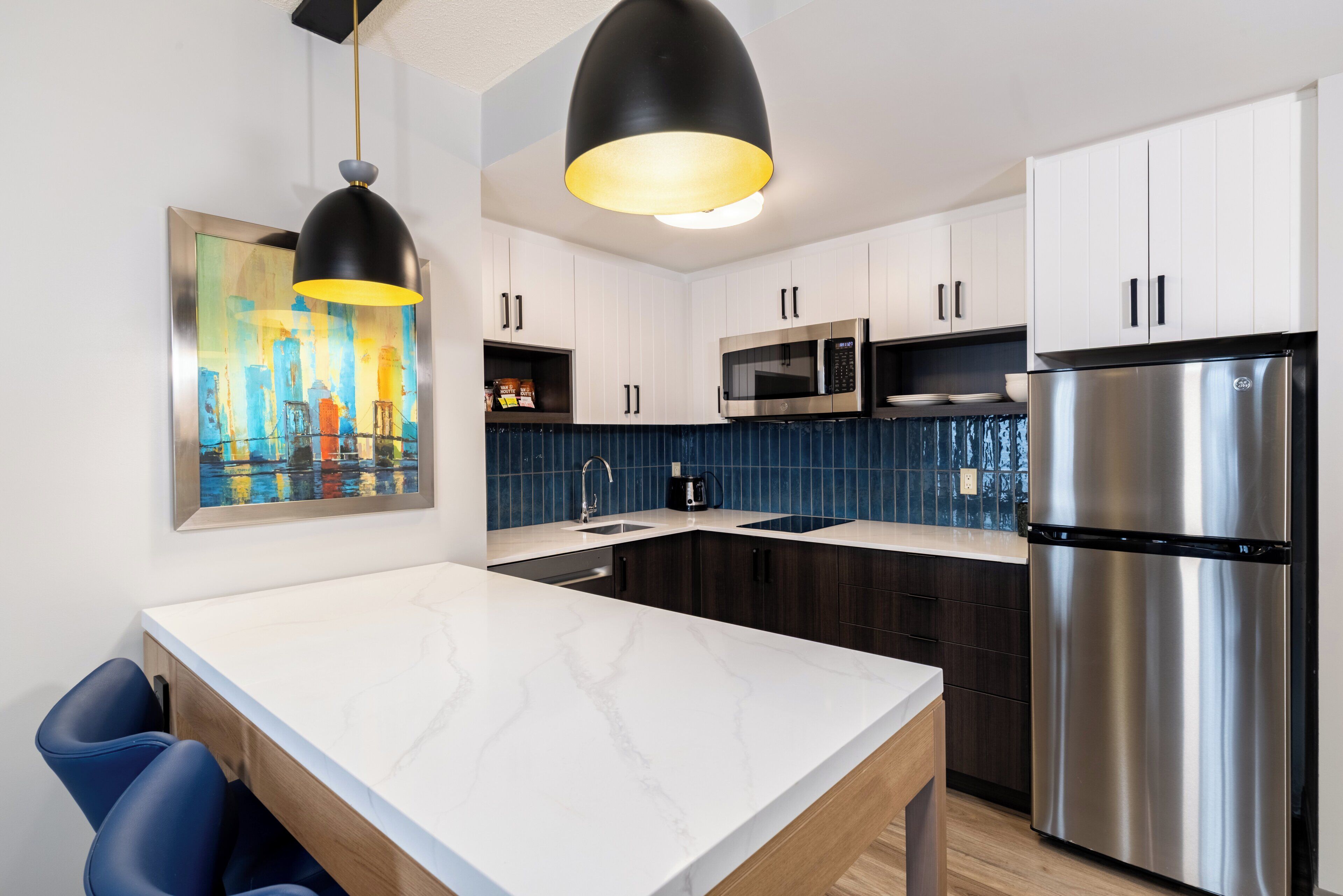 A modern kitchen with white cabinets, a stainless steel refrigerator, a microwave, and a large island with blue chairs.