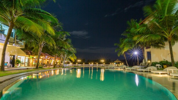 A nighttime view of a swimming pool with palm trees and a building with lights. - River Beach Resort and Residences (Da Nang)