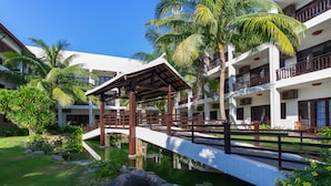 A bridge over a pond, surrounded by lush greenery and palm trees, leading to a building with balconies and a white facade. - River Beach Resort and Residences (Da Nang)