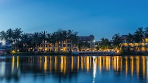A waterfront hotel with multiple buildings, palm trees, and a calm body of water reflecting the lights. - River Beach Resort and Residences (Da Nang)