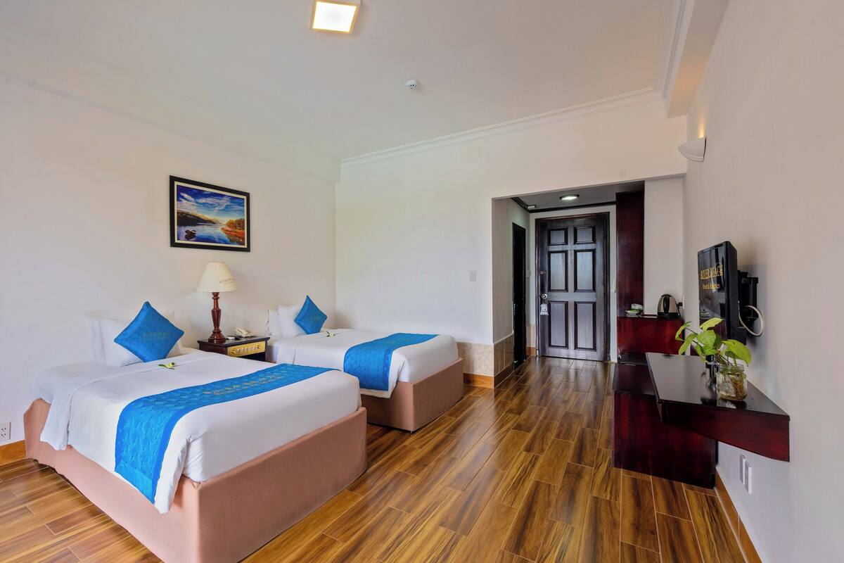 A hotel room with two beds, a desk, a chair, a lamp, a painting, and a door leading to another room.