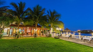 An outdoor restaurant with tables and chairs, palm trees, and a view of the water. - River Beach Resort and Residences (Da Nang)