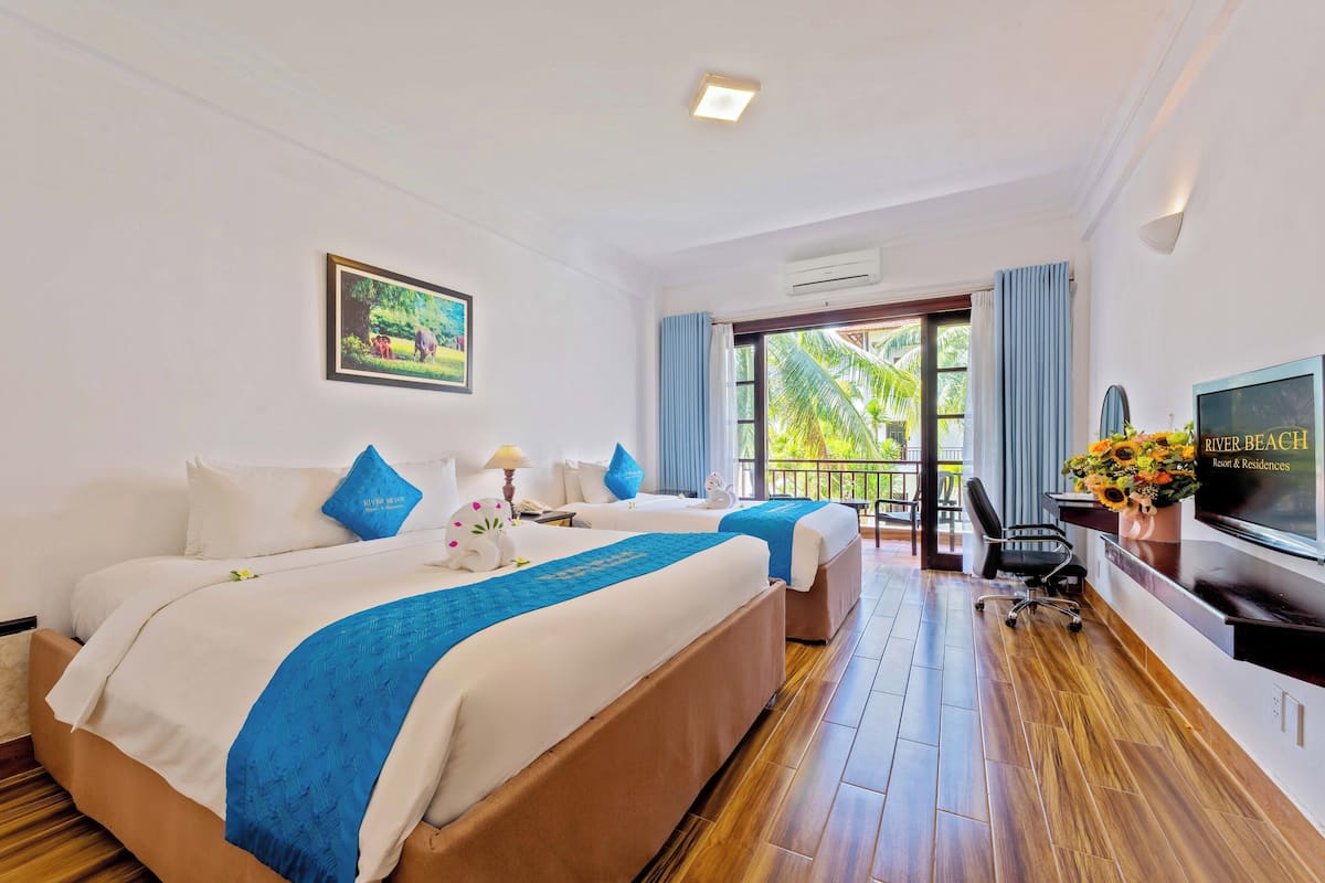 A hotel room with two beds, a TV, a desk, and a balcony with a view of palm trees.