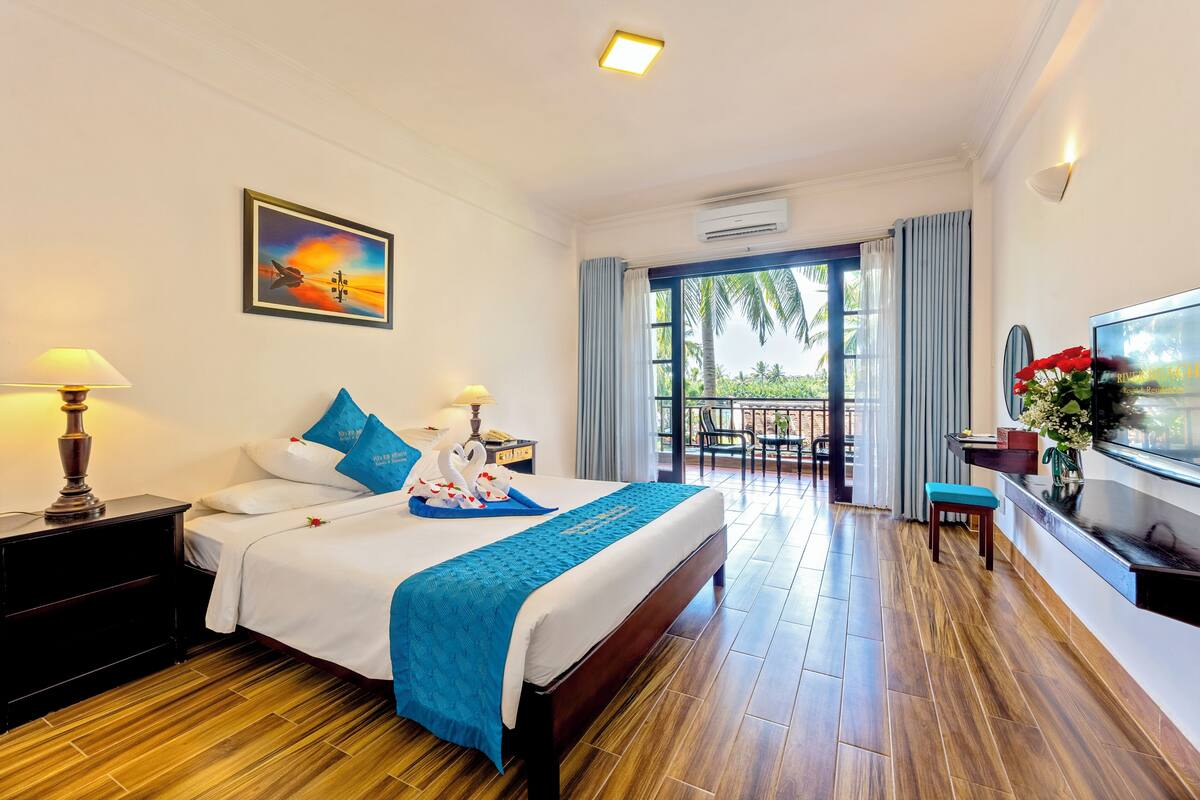 A hotel room with a large bed, a TV, a desk with a chair, and a balcony with a view of palm trees.