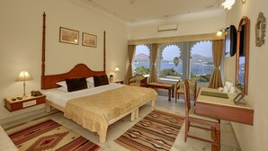 A hotel room with a large bed, a desk, a chair, a nightstand, a lamp, a mirror, and a view of the sea. - Rampratap Palace By Fateh Collection (Udaipur)