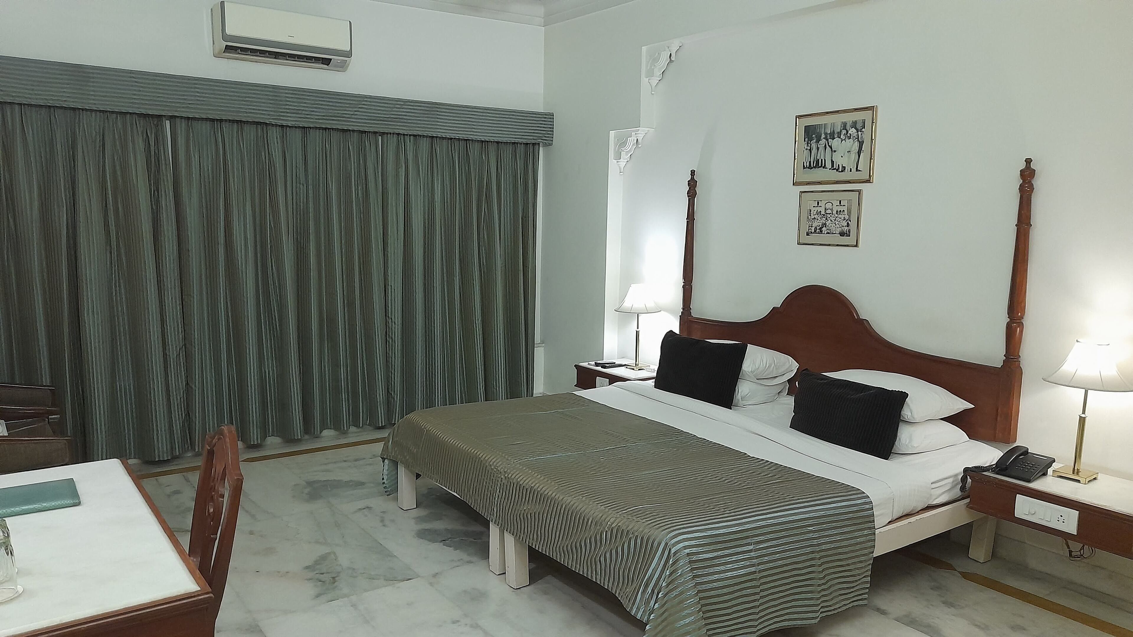 Habitación de hotel con cama, mesitas de noche, escritorio, silla, ventana con cortinas y aire acondicionado instalado en la pared.
