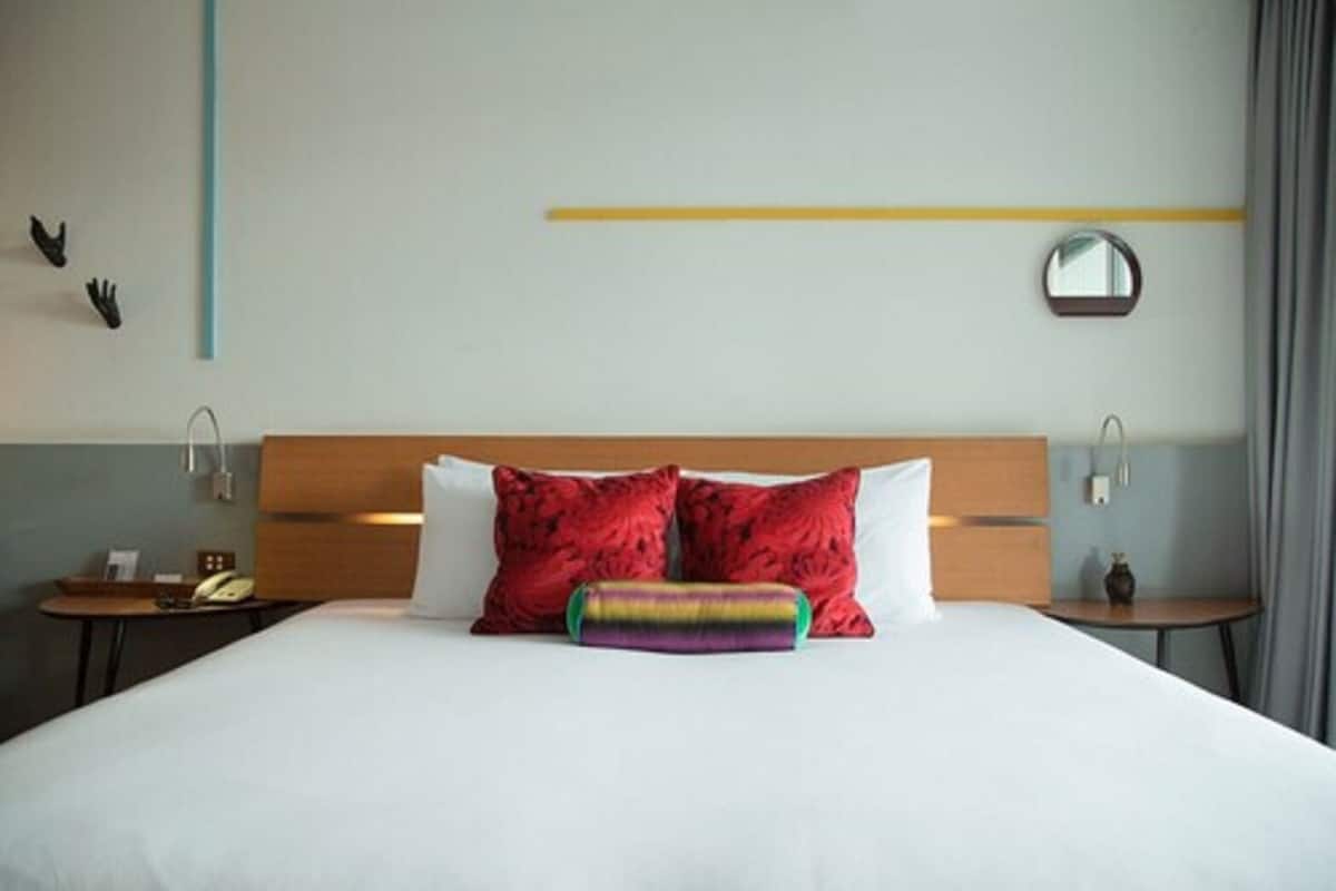 A hotel room with a large bed, two red pillows, a colorful pillow, a bedside table with a lamp, a small table with a vase, and a mirror on the wall.