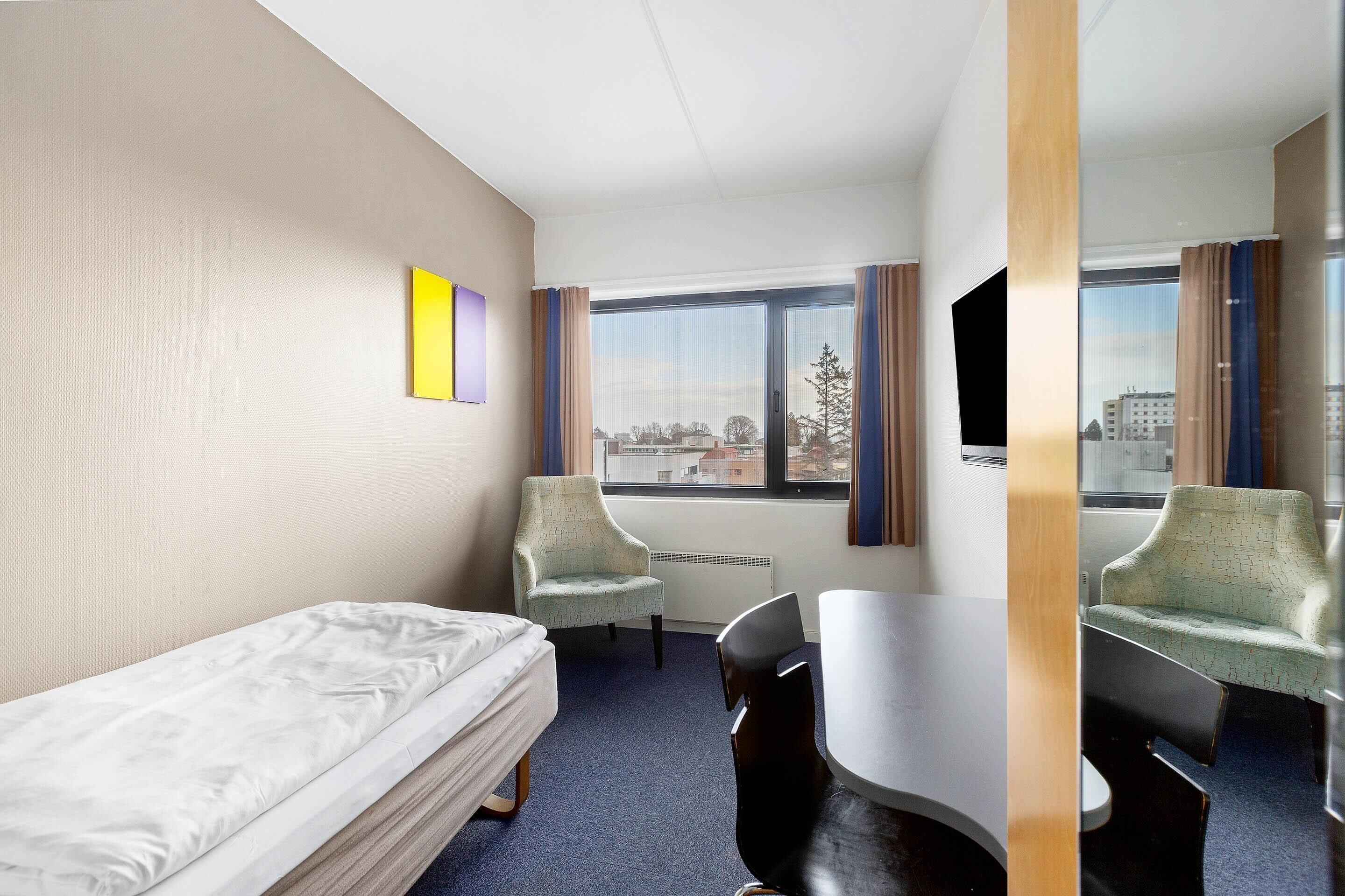 Habitación de hotel con cama, dos sillas, una mesa, televisor y ventana con vista a la ciudad.