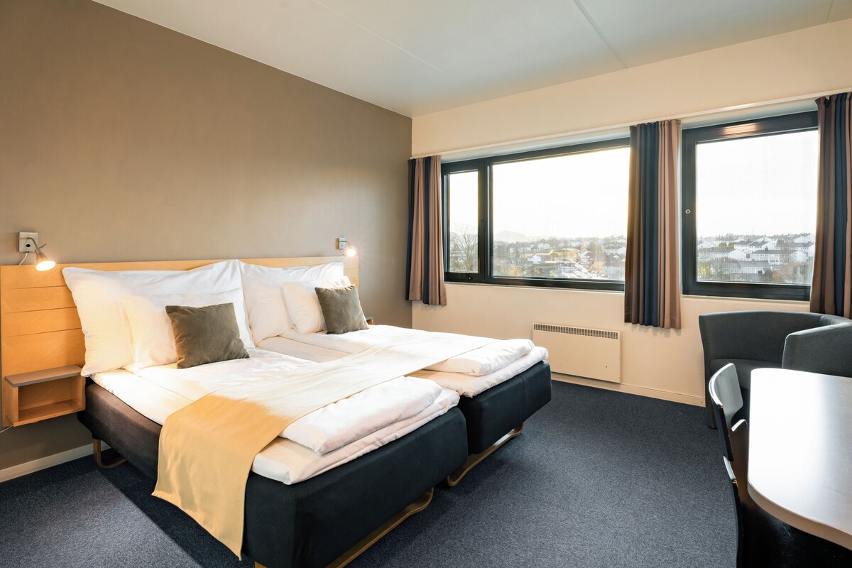 A hotel room with a bed, bedside table, chair, and a large window with curtains.