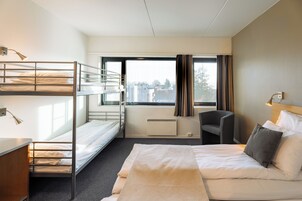 A hotel room with bunk beds, a single bed, a desk, a chair, and a large window with curtains.