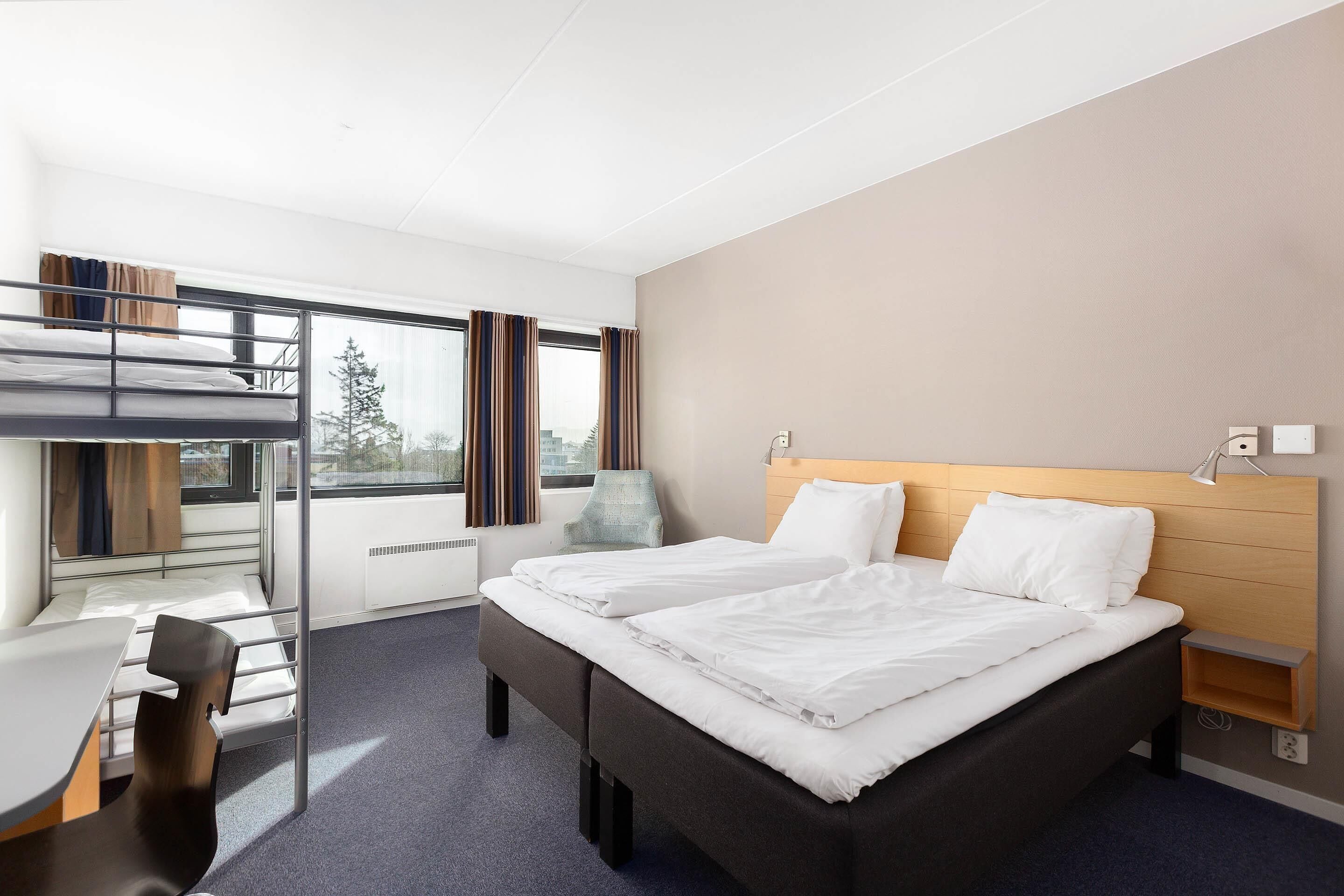 A hotel room with a bunk bed, a desk, a chair, a bedside table, and a window with curtains.