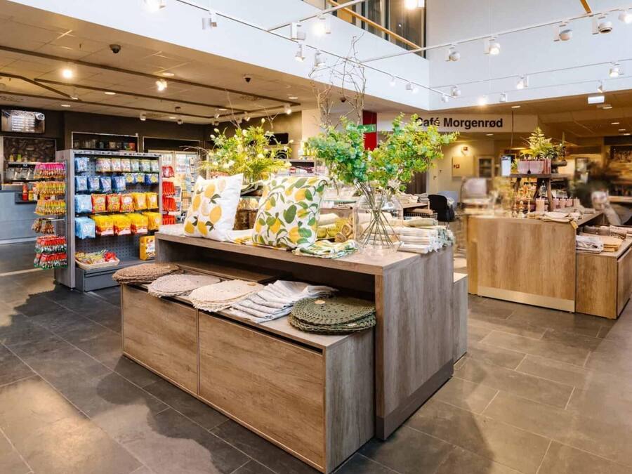 Un intérieur moderne avec un espace d’exposition central où sont présentés des textiles et des plantes, un café en arrière-plan et un comptoir de caisse.