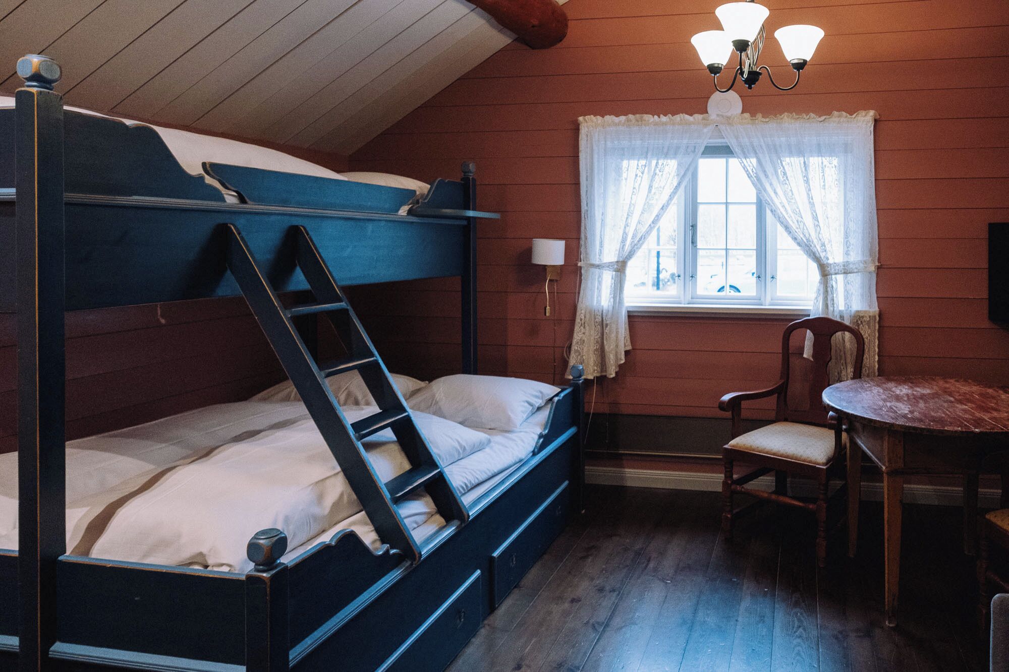 A bunk bed with a ladder, a small table, and a chair in a room with wooden walls and a window with lace curtains.