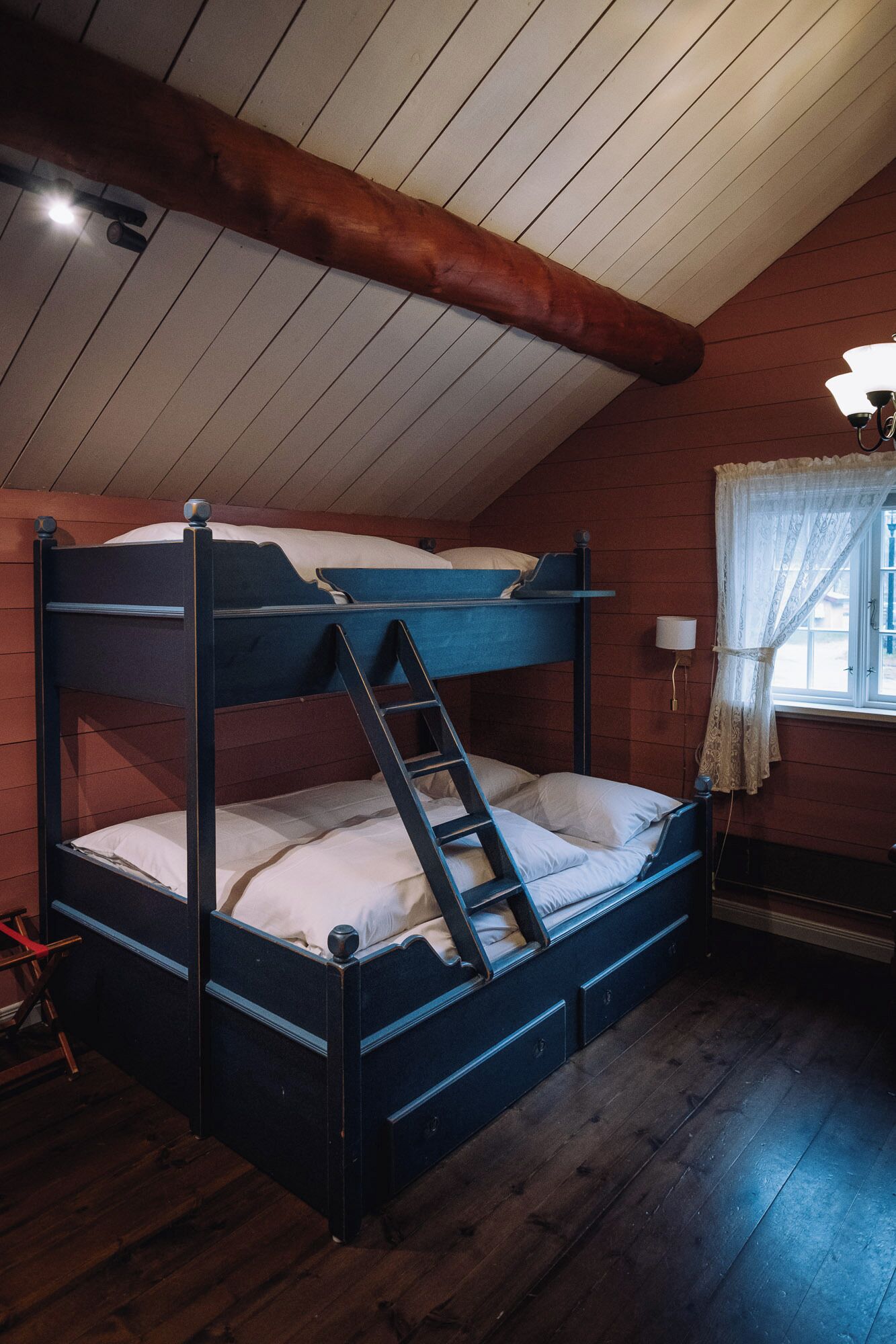 A bunk bed with a ladder, wooden walls, a window with curtains, and a wooden floor.