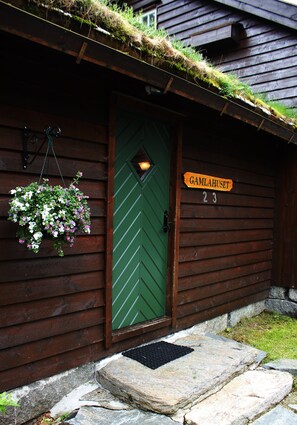 Una cabaña de madera con una puerta verde, una cesta de flores colgante y un letrero que dice "GAMLAHUSET 23".