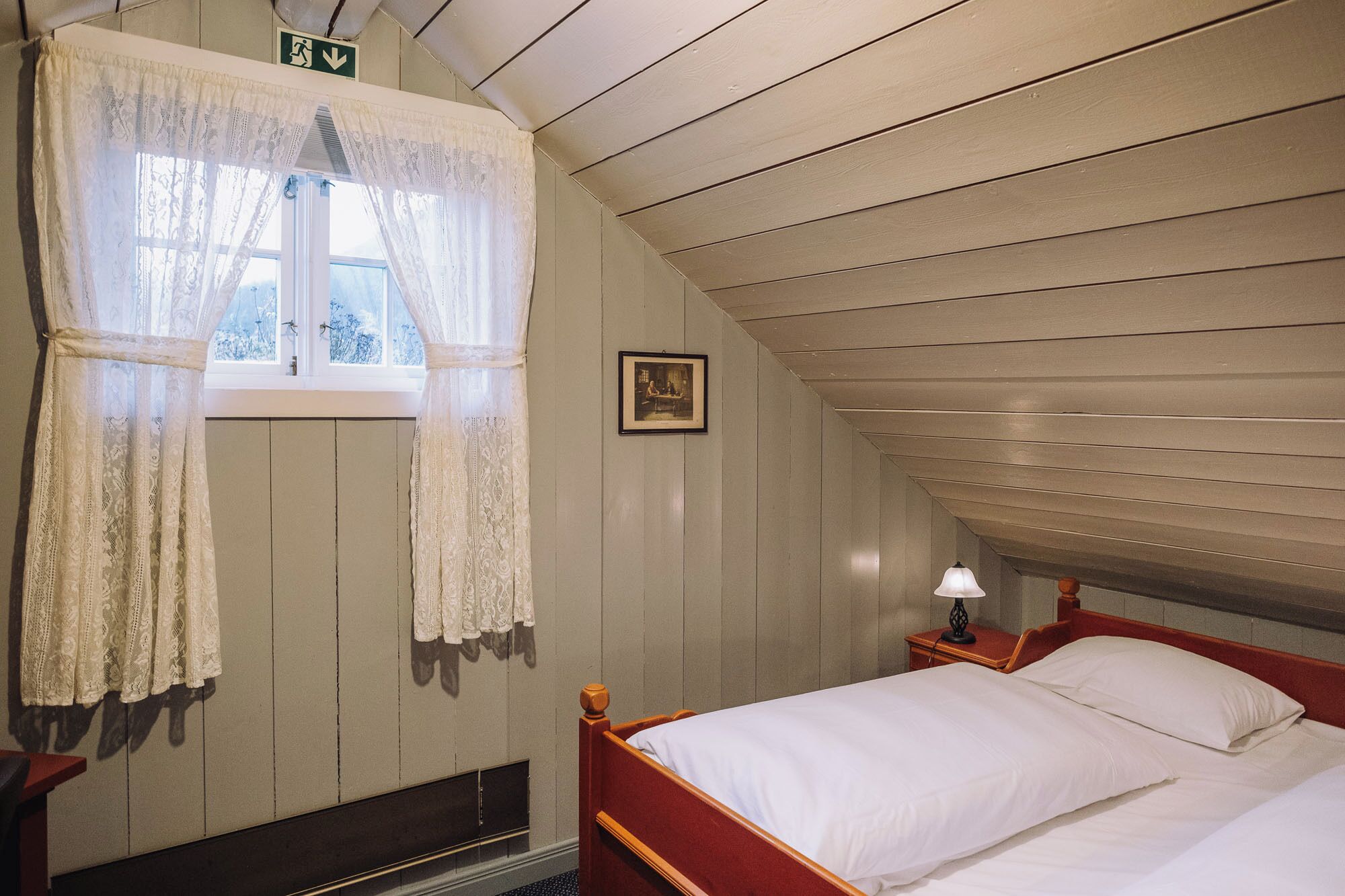 Ein Schlafzimmer mit Holzbalkendecke, einem Einzelbett, einem Nachttisch mit Lampe, einem Fenster mit Spitzenvorhängen und einem gerahmten Bild an der Wand.