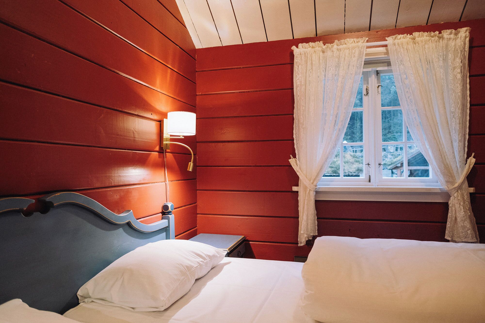 Una habitación con pared de madera, una cama con ropa blanca, una mesita de noche, una lámpara y una ventana con cortinas de encaje.
