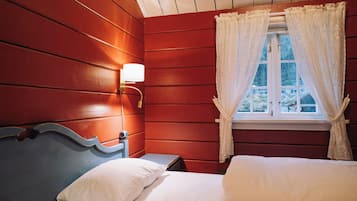 Een kamer met een houten wand, een bed met wit beddengoed, een nachtkastje, een lamp en een raam met kantgordijnen.