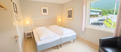 A double bed with white linens, a bedside table, a lamp, and a window with a view of a parking lot and mountains.