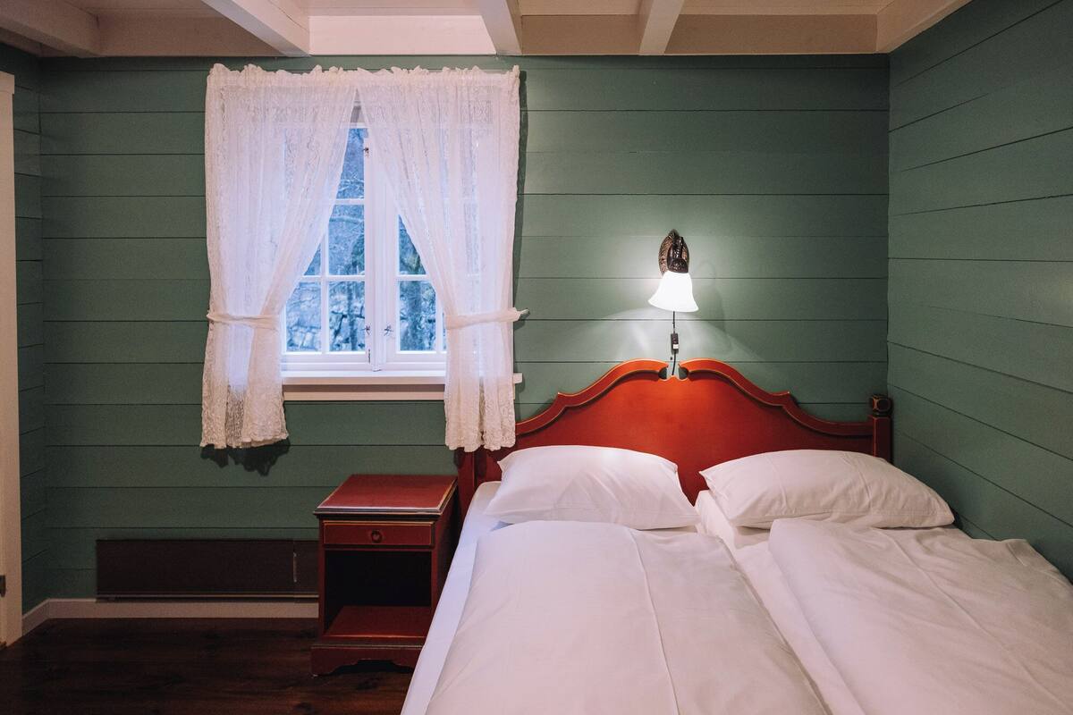 A room with a bed, a nightstand, a window with lace curtains, and a wall-mounted lamp.