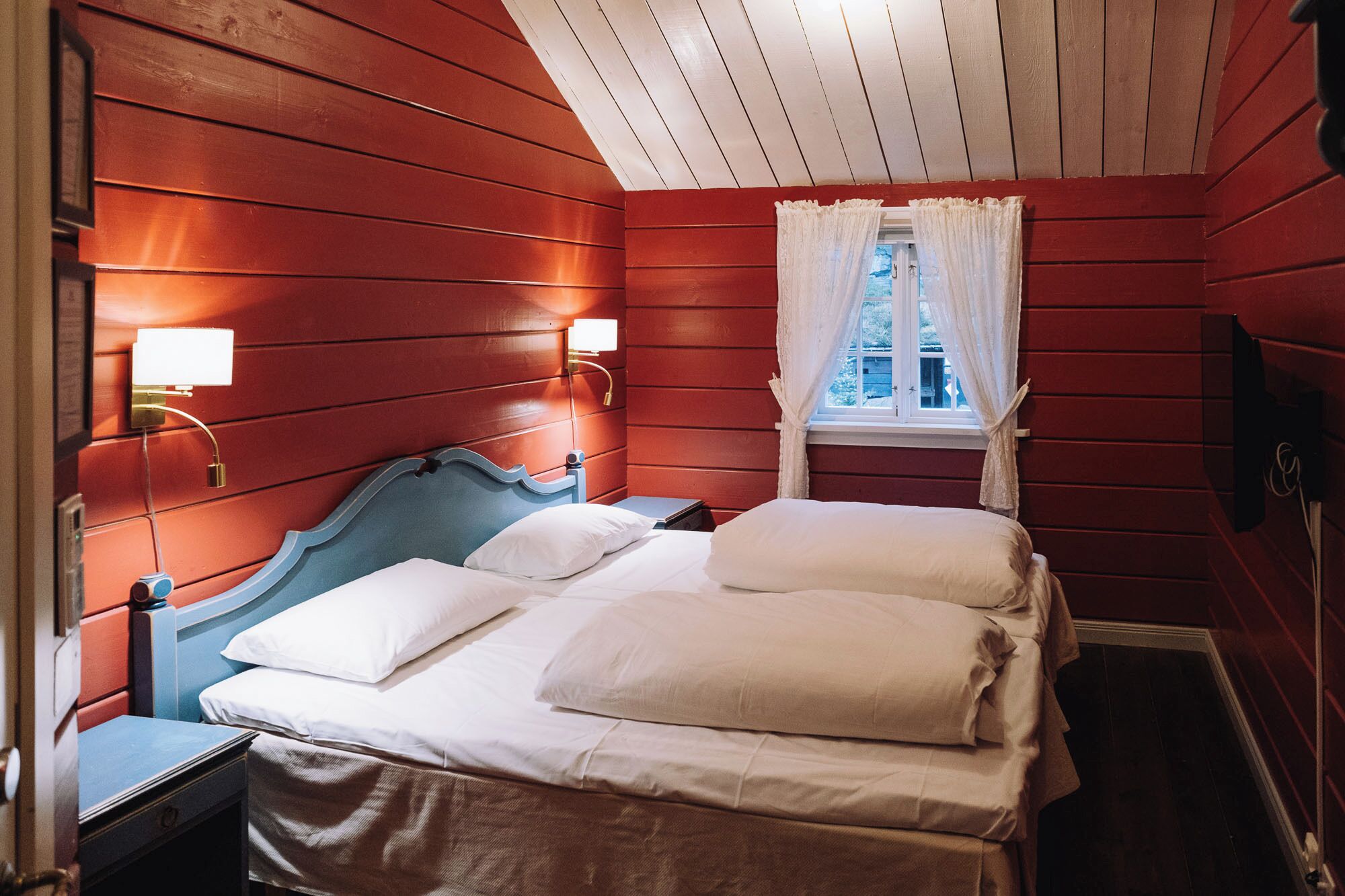 Quarto com cama, mesa de cabeceira, janela com cortinas brancas e luminárias fixadas na parede.