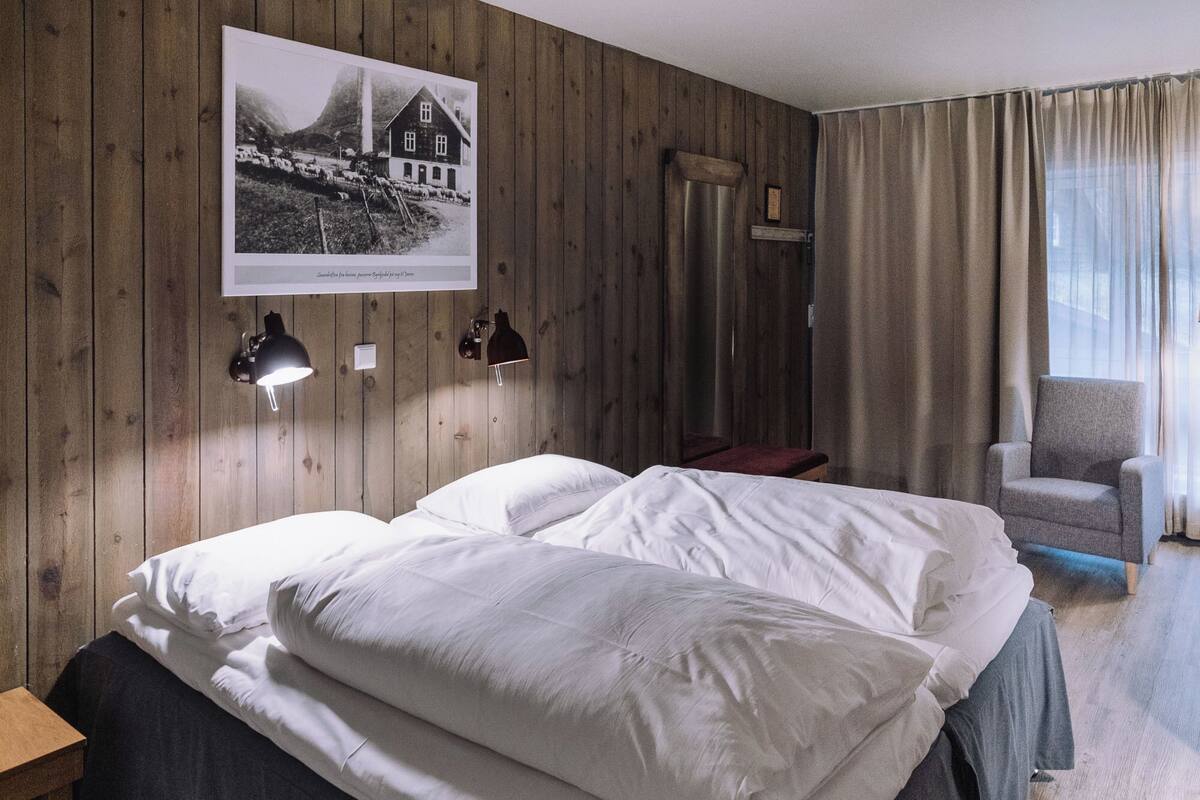 A hotel room with wooden panel walls, a bed with white linens, a chair, and a window with curtains.