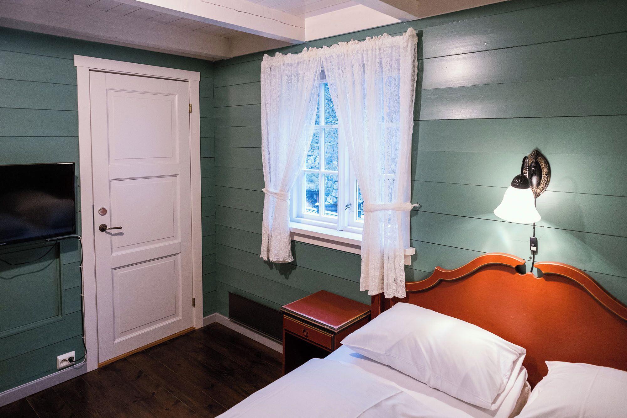 A room with a bed, a nightstand, a lamp, a television, and a window with curtains.