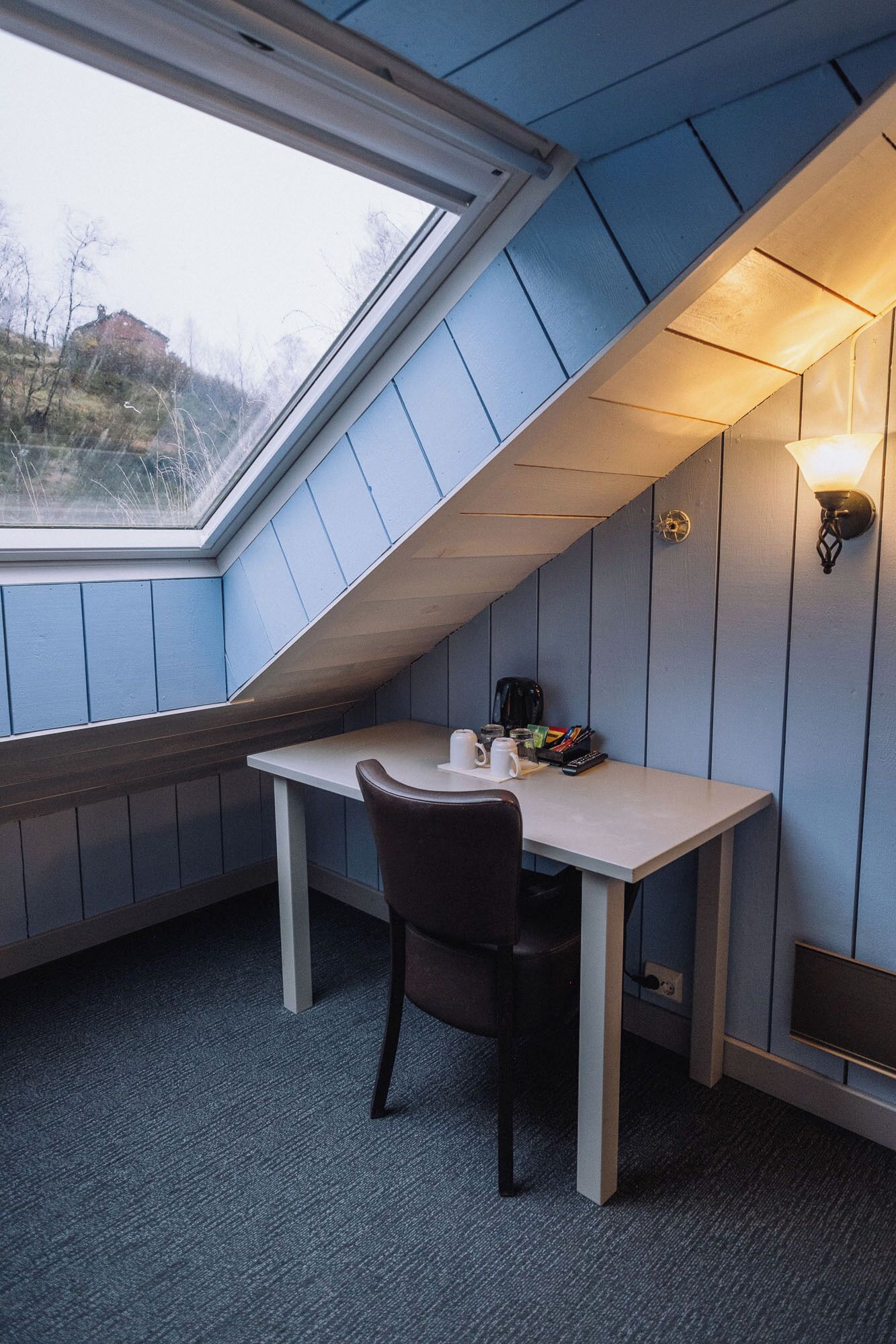 A small, sloped room with a skylight, a desk, a chair, and a wall-mounted light fixture.