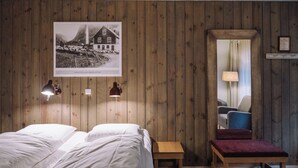 Una habitación con paneles de madera, una cama con sábanas blancas, un pequeño banco de madera, un espejo y un cuadro en la pared.