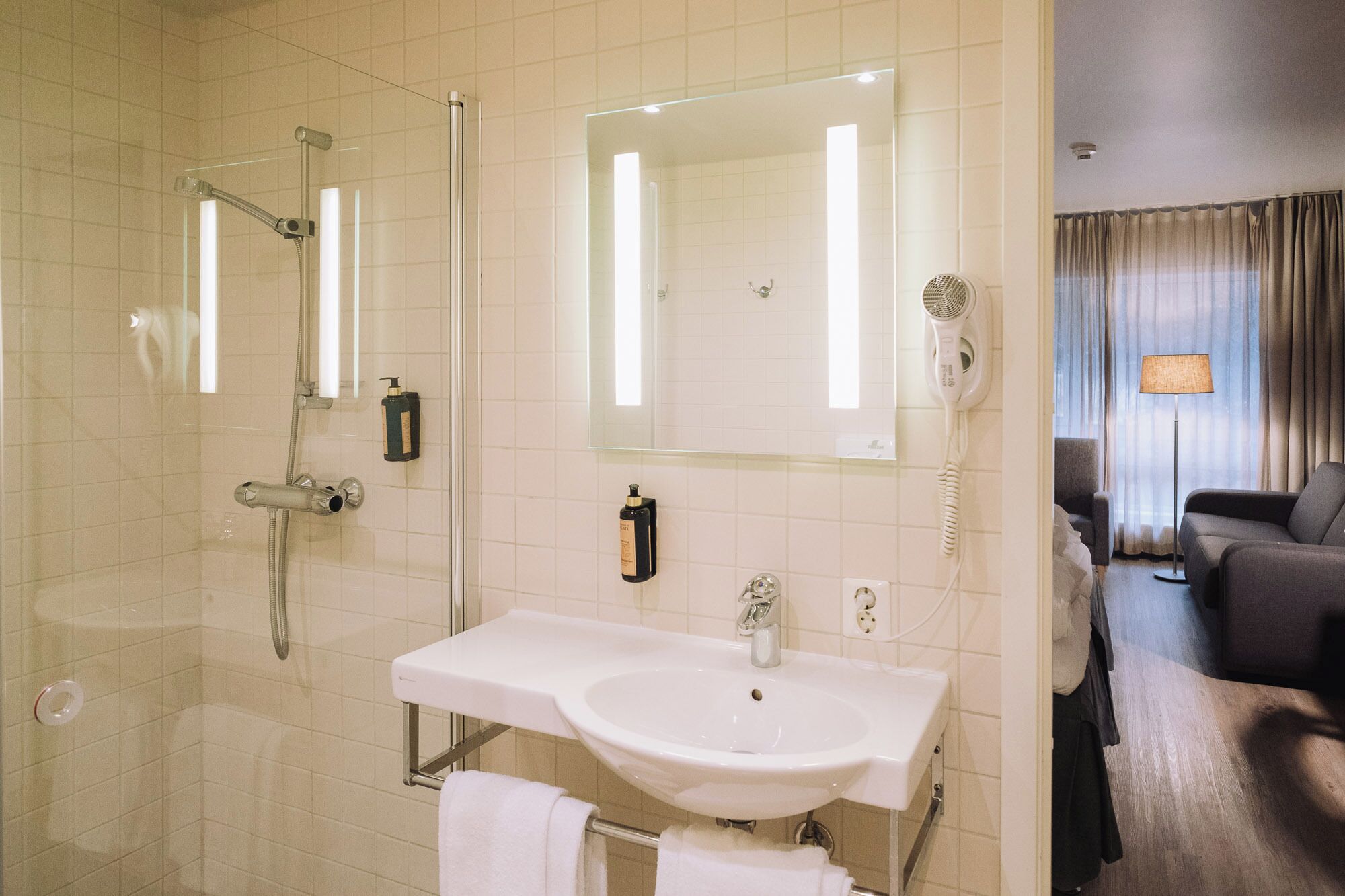 A hotel bathroom with a shower, sink, and mirror. There is a hairdryer and a towel rack.