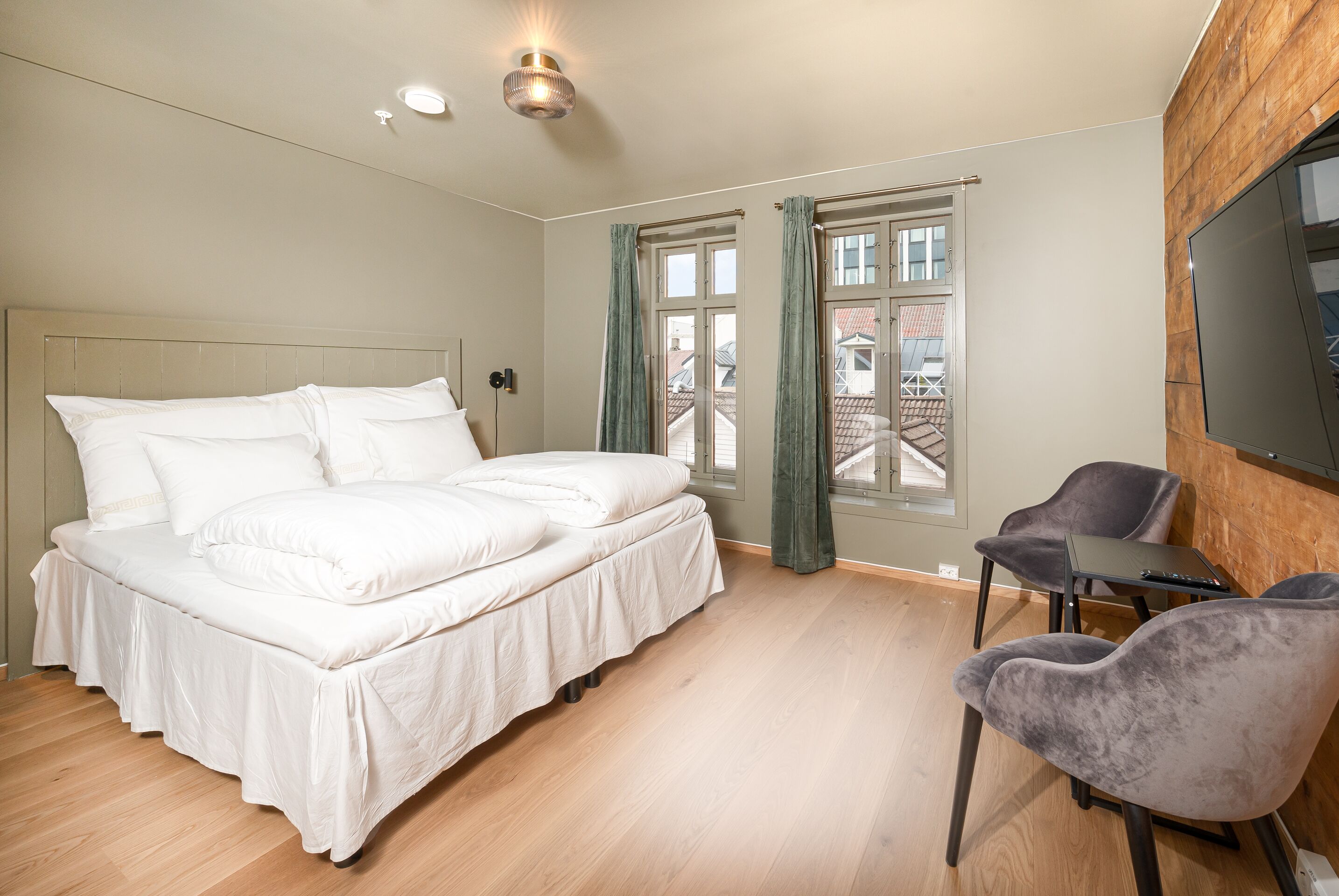 Une chambre avec un grand lit, deux fenêtres avec des rideaux, un téléviseur fixé au mur et une petite table avec des chaises.